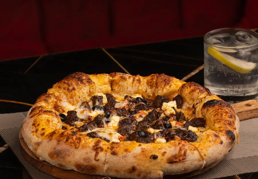 Pizza boulfaf à la croûte soufflée dorée, merguez et olives, sur planche de bois, verre citronné à côté