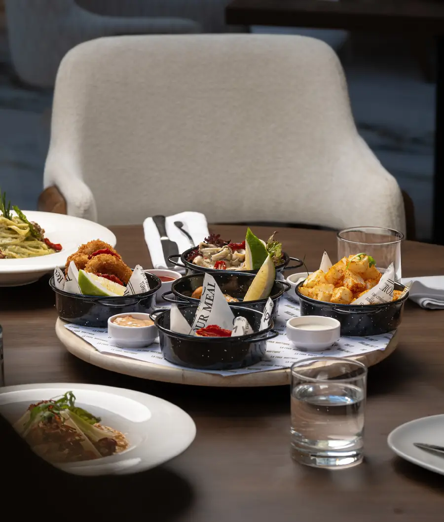 Plateau de cassolettes noires : calamars panés, pommes de terre épicées, sauces, sur table ronde en bois