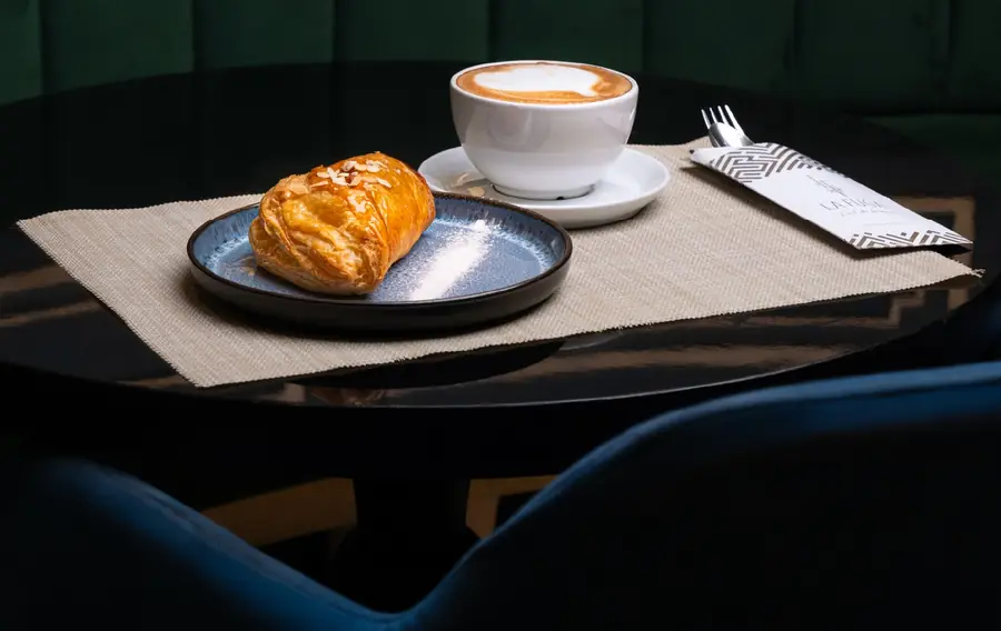Un cappuccino et une viennoiserie aux amandes sur la table laquée noire, banquette de velours émeraude derrière