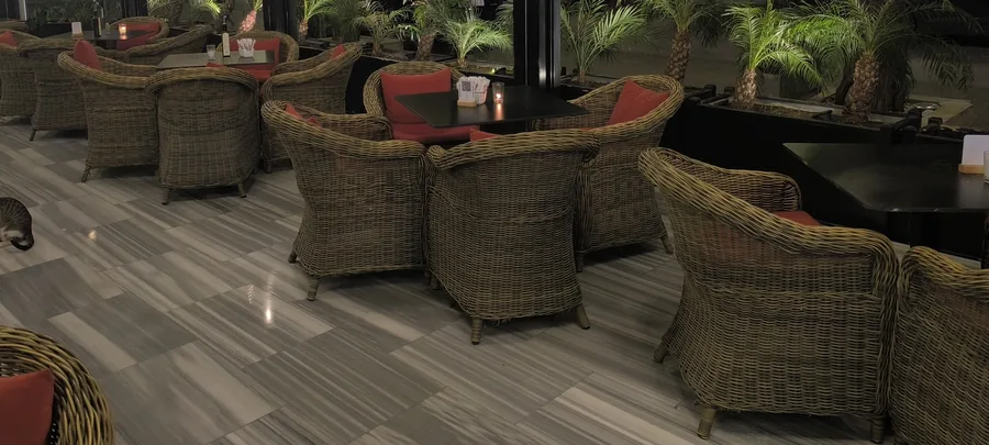 La terrasse couverte le soir : fauteuils en rotin aux coussins rouges, palmiers, sol de marbre gris
